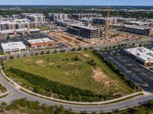 Aerial view of a construction site with a multi-story building under construction and a crane, adjacent to a commercial complex and a green, undeveloped lot nearby.