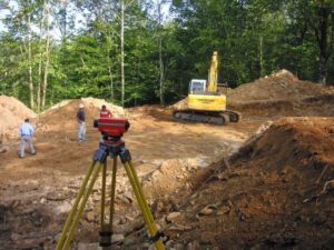 Surveying setup on a construction site: a red instrument on a yellow tripod foreground, excavator and dirt piles in background.